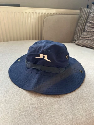 J.Lindeberg bucket hat / golfhatt mörkblå - Mörkblå J.Lindeberg-hatt med bred skärm och klassisk JL-logga framtill. Praktisk modell med ventilationsdetaljer, justerbart hakband och tryckknappar på sidorna så att skärmen kan fästas upp. Perfekt för golf, fiske, vandring eller andra aktiviteter utomhus.  Fint begagnat skick med normala mindre spår av användning.  