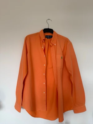 Orange skjorta från Ralph Lauren - Säljer en orange skjorta från Ralph Lauren med klassisk krage och knappstängning framtill. Skjortan har lång ärm och en liten grön logga broderad på bröstfickan. Perfekt för dig som vill sticka ut med färg!