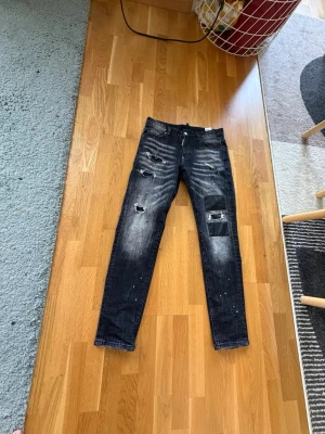 jeans från Dsquared2 - Säljer ett par svarta jeans från Dsquared2 med slitna detaljer, hål och patch på ena benet. Jeansen har en smal passform och cool tvätt med vita färgstänk. Perfekta för dig som gillar streetwear och edgy stil.