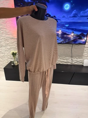 Randigt beige loungewear-set - Mysigt loungewear-set i beige med tunna vita ränder. Setet består av en långärmad tröja med rund hals och avslappnad passform samt matchande byxor med dragsko i midjan. Perfekt för chill dagar hemma eller när du vill vara bekväm men ändå snygg.