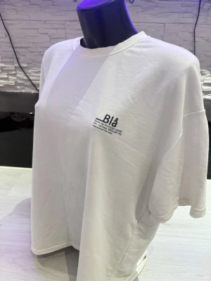 Vit oversized t-shirt med tryck - Vit oversized t-shirt med svart texttryck på bröstet där det står 'Blå' och ett citat om havet och himlen. T-shirten har rund hals och är tillverkad i mjuk bomull. Perfekt för en avslappnad och trendig stil.