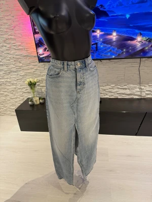 Ljusblå lång jeanskjol med slits - Trendig ljusblå lång jeanskjol i klassisk denim med hög midja och rå kant nertill. Kjolen har en tydlig slits framtill och bakfickor. Perfekt för dig som gillar en avslappnad men snygg stil.