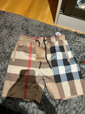 Burberry rutiga badbyxor - Säljer ett par klassiska badbyxor från Burberry med det ikoniska rutmönstret i beige, svart, vitt och rött. Modellen har elastisk midja med dragsko och sidofickor. Perfekta för stranden eller poolen och ger en lyxig känsla till din sommarstil.