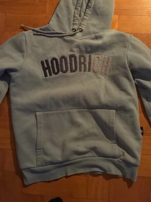 Ljusblå hoodie från Hoodrich - Snygg ljusblå hoodie från Hoodrich med stor magficka och broderad logga på bröstet. Klassisk huva med dragsko och mjukt material som känns skönt mot huden. Perfekt för en avslappnad streetwear-look.