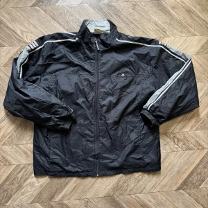 Svart vindjacka från Adidas - Snygg svart vindjacka från Adidas med vita och grå detaljer längs ärmarna och axlarna. Jackan har dragkedja framtill, ståkrage och elastiska muddar vid ärmsluten. Klassisk Adidas-logga på bröstet och ryggen. Perfekt för en sportig och avslappnad stil.