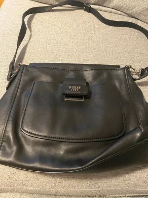 Svart handväska från Guess - Snygg svart handväska från Guess med justerbar axelrem och stor ficka framtill. Väskan har ett lock med metallspänne där det står 'Guess 1981'. Tillverkad i slätt konstläder och har en stilren, modern design.