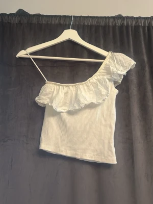 Vit oneshoulder topp med volang - Säljer en söt vit oneshoulder topp med volangdetalj längs halsringningen. Toppen är ribbad och har en smal axelrem på ena sidan. Perfekt för sommarens alla tillfällen!