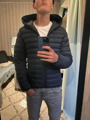 Mörkblå save the duck jacka - Säljer en stilren mörkblå dunjacka med huva och horisontella sömmar. Jackan har dragkedja framtill och en smal passform som ger en modern look. Perfekt för kalla vinterdagar och enkel att matcha med olika outfits.