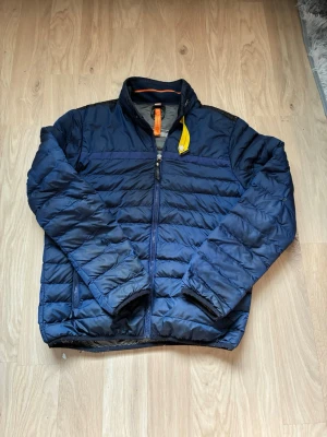 Mörkblå pufferjacka med gul detalj  - Säljer en mörkblå pufferjacka med dragkedja och hög krage. Jackan har en gul knappdetalj vid halsen och orange insida vid kragen. Quiltad design och två sidofickor. Perfekt för kalla dagar och riktigt skön att bära.
