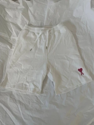 Vita shorts med snörning och hjärtdetalj - Snygga vita shorts med snörning i midjan och en röd hjärtdetalj med bokstaven A på ena benet. Mjukt material och elastisk midja för skön passform. Perfekta för varma dagar och chill outfits.