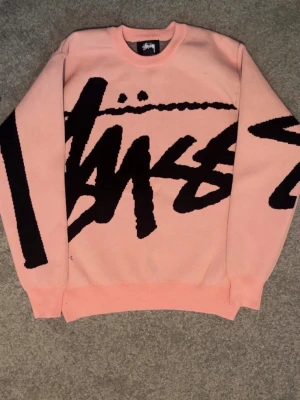 Rosa stickad tröja från Stüssy - Säljer en rosa stickad tröja från Stüssy med stort svart grafiskt tryck över hela framsidan och ena ärmen. Tröjan har rund halsringning och ribbade muddar vid ärmslut och nederkant. Perfekt för dig som gillar streetwear och vill sticka ut.