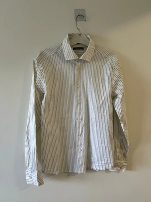 Vit randig skjorta från Mansfield - Klassisk vit skjorta från Mansfield med tunna svarta ränder. Skjortan har lång ärm, knappar framtill och en stilren krage. Perfekt för en clean och snygg look. Tillverkad i bomull för skön känsla.