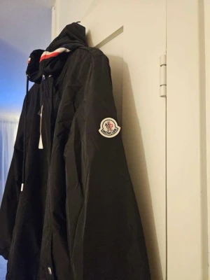 Svart Moncler windbreaker med huva - Snygg svart vindjacka från Moncler med dragkedja framtill, två fickor och justerbar huva med vita och röda detaljer. Jackan har Moncler-logga på ärmen och elastiska muddar vid ärmslut och nederkant. Perfekt för blåsiga dagar och streetwear-stil.