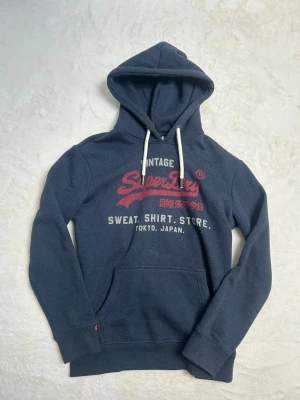 Mörkblå Superdry hoodie med tryck - Mörkblå hoodie från Superdry med stor huva, vita dragsnören och känguruficka. Tryck i rött och vitt framtill med texten 'Vintage Superdry Sweat. Shirt. Store. Tokyo, Japan.' Skön och avslappnad passform, perfekt till vardags.