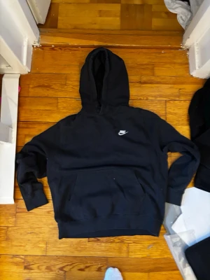Svart hoodie från Nike med logga - Svart hoodie från Nike med klassisk vit logga på bröstet. Hoodien har stor huva och en rymlig magficka framtill. Tillverkad i mjukt material och har ribbade muddar vid ärmslut och nederkant. Perfekt för en avslappnad och sportig stil.