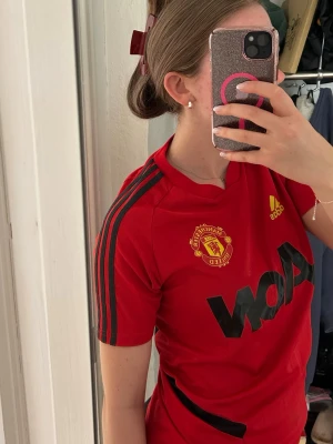Manchester United röd Adidas tröja - Säljer en röd Manchester United träningströja från Adidas med svarta detaljer och klubbmärke på bröstet. Tröjan har korta ärmar, svarta ränder på axlarna och stor svart AON-logga framtill. Perfekt för fotbollsträning eller som supporterplagg.
