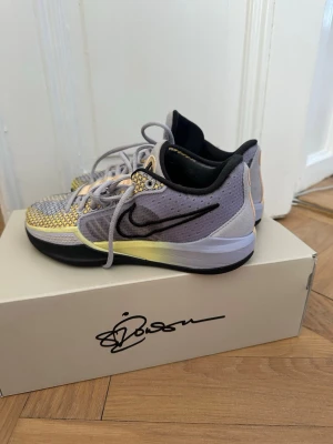 Sabrina 1 basketsko - Snygga basketskor från Nike ”Sabrina 1” grått och gult med svart swoosh på sidan. Ovandelen har ett mönstrat meshmaterial och detaljer i beige. Skorna har lågt skaft och svart sula. Perfekta för dig som vill sticka ut med coola färgkombinationer.