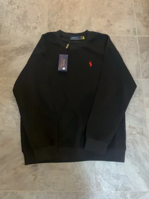 Svart stickad tröja Polo Ralph Lauren - Snygg svart stickad tröja från Polo Ralph Lauren med klassisk röd logga broderad på bröstet. Tröjan har rund halsringning och ribbade muddar vid ärmslut och nederkant. Perfekt för en stilren och enkel look tröjan passar både L och M den säljs eftersom fick den i present men tyvärr så passade den inte mig därför vill jag sälja den för en riktigt billig pris och den är endast testad hemma