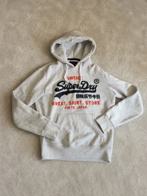 Ljusgrå Superdry hoodie med tryck - Snygg ljusgrå hoodie från Superdry med stor logga och text i svart och rött på bröstet. Klassisk huva med dragsko och magficka framtill. Tillverkad i mjukt sweatshirtmaterial, perfekt för en avslappnad stil.