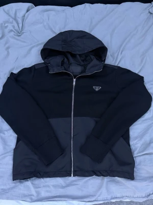 Prada jacka  - Snygg svart hoodie från Prada med dragkedja framtill och ikonisk trekantig logga på bröstet. Hoodien har en huva och är tillverkad i en mix av stickat tyg och glansigt material på huva och nederdel. Perfekt för en stilren och modern look.