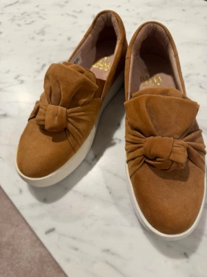 Bruna sneakers med rosett och platåsula - Snygga bruna sneakers i mockaimitation med stor dekorativ rosett på ovansidan. Skorna har en vit platåsula och slip-on design utan snörning. Perfekta för dig som vill ha något unikt och trendigt till vardagslooken.