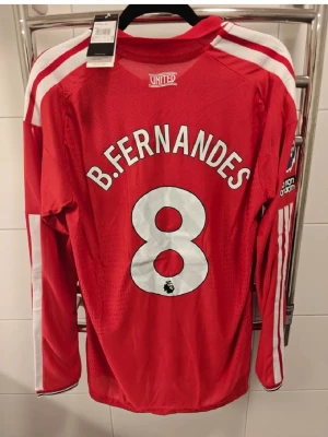 Manchester United spelar version långärmad matchtröja - Säljer en röd långärmad Manchester United matchtröja med B. Fernandes och nummer 8 på ryggen. Tröjan har vita detaljer, klubbmärke och Adidas-logga på bröstet samt Premier League-märke på ärmen. Perfekt för fotbollsfans!
