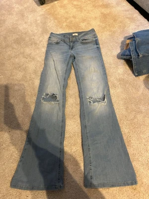 Ljusblå bootcut jeans med slitningar - Säljer ett par ljusblå lågmidjade bootcut jeans med slitna hål på båda knäna. Jeansen är ifrån Nelly och är i storlek 36. De är stretchiga i materialet