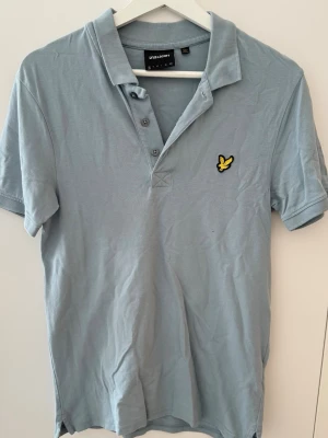 Ljusblå pikétröja från Lyle & Scott - Klassisk kortärmad pikétröja från Lyle & Scott i en stilren ljusblå / duvblå nyans. Tröjan har vikbar krage, knappknäppning framtill och den ikoniska gula örn-logotypen broderad på bröstet. Tillverkad i skön bomullskvalitet med fin struktur. OBS: Ett mycket litet märke/skavank finns nedanför logotypen på framsidan (syns på närbilden).
