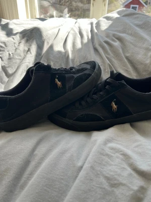 Svarta sneakers från Polo Ralph Lauren - Snygga svarta sneakers från Polo Ralph Lauren med klassisk låg profil. Skorna har svart mockadetalj och tyg, samt den ikoniska broderade loggan i guld på sidan. Perfekta för dig som gillar stilrena och tidlösa sneakers med en sportig touch.