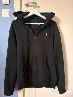 Svart hoodie från Polo Ralph Lauren - Svart hoodie med dragkedja från Polo Ralph Lauren. Klassisk design med huva och dragsnören, fickor framtill och liten gul broderad logga på bröstet. Insidan av huvan har ett rutigt mönster i grönt och blått.