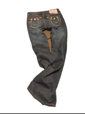 True Religion mörkblå jeans - Säljer ett par mörkblå jeans från True Religion med ikoniska kontrastsömmar i gult och orange. Baksidan har stora fickor med dekorativa broderier och knappar. Klassisk femficksmodell i denim med raka ben och låg midja. midjemått 36cm innerbenslängd 78cm
