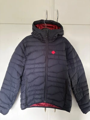 Svart pufferjacka från J.Lindeberg - Snygg svart pufferjacka från J.Lindeberg med röd innerfoder och huva. Jackan har två dragkedjeförsedda fickor framtill och en diskret logga på bröstet. Perfekt för kalla dagar när du vill hålla värmen och ändå se stilren ut. Väldigt bra skick 