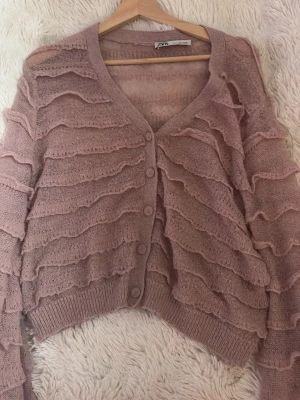 Rosa kofta med volangdetaljer - Superfin rosa kofta med volangliknande ränder över hela plagget. Koftan har v-ringning, knäppning framtill och ribbade muddar vid ärmslut och nederkant. Perfekt för dig som gillar en söt och trendig stil.