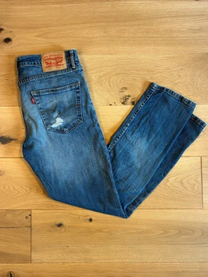 Levi's blå raka jeans - Klassiska Levi’s 502-jeans i medelblå tvätt med naturligt slitna detaljer. Modellen är rak/regular fit och har en snygg, äkta använd look med fin whiskering vid benen.  •	Modell: Levi’s 502 	•	Storlek: W34 L32 	•	Skick: Använt skick med charmigt slitage - bl.a. ett litet hål/nött parti på bakfickan   	•	Färg: Medelblå denim med fin tvätt och naturliga fadingar 	•	Äkta läderlapp och röd Levi’s-etikett på bakfickan