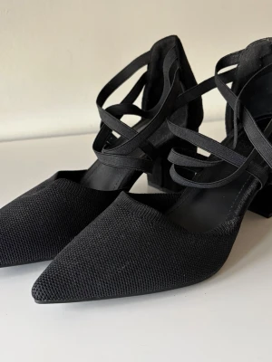 Svarta pumps med blockklack och band - Snygga svarta pumps med spetsig tå, bred 7cm blockklack och korsade band över foten. Ovandelen är i textil och klacken i mockaimitation. Perfekta för dig som vill ha både stil och komfort. Oerhört sparsamt använda så är i topp/nyskick! Ärliga och snabba affärer uppskattas:)