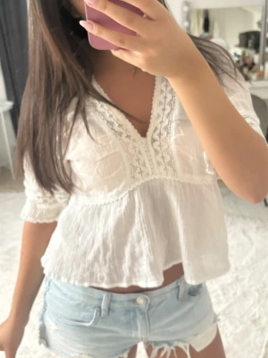 Vit blus - Säljer en luftig vit blus i boho-stil med v-ringning och fina spetsdetaljer längs halsringningen. Blusen har trekvartsärm och är gjord i ett tunt, skönt material som ger en avslappnad look. Perfekt till jeansshorts eller kjol.