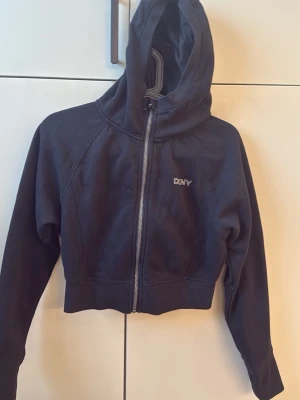 Svart cropped hoodie från DKNY Sport - Snygg svart cropped hoodie från DKNY Sport med dragkedja framtill och liten logga på bröstet. Hoodien har långa ärmar och bred mudd i nederkant. Perfekt för en sportig och avslappnad stil. Tillverkad i mjukt material för extra komfort.