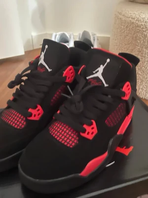 Svarta och röda Air Jordan 4 sneakers - Säljer ett par Air Jordan 4 sneakers i svart och rött med vita detaljer. Skorna har en hög siluett, meshpaneler och klassisk Jumpman-logga på plösen och hälen. Materialet är en mix av mocka och syntet. Perfekta för dig som vill sticka ut med din stil.