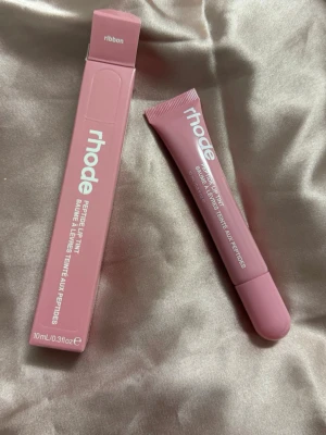 rhode Peptide Lip Tint Ribbon - Säljer ett rhode Peptide Lip Tint i färgen Ribbon. Läppbalsamet kommer i en mjuk rosa tub med vit text och matchande förpackning. Innehåller 10 ml och har en smidig applikator. Perfekt för dig som vill ha återfuktade och lätt färgade läppar.