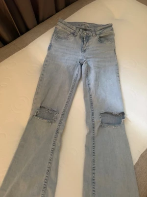 Ljusblå slitna bootcut jeans - Säljer ett par ljusblå bootcut jeans med slitningar över knäna och broderade detaljer på bakfickorna. Jeansen har klassisk femficksdesign och är gjorda i mjukt denimtyg. Perfekta för en avslappnad och trendig look.