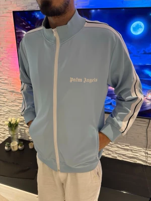 Ljusblå track jacket från Palm Angels - Säljer en ljusblå track jacket från Palm Angels med vita och svarta ränder längs ärmarna. Jackan har hög krage, hel dragkedja framtill och ribbade muddar. Perfekt för en sportig och avslappnad stil.