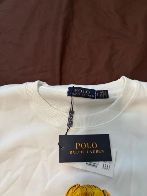 Vit sweatshirt Polo Ralph Lauren - Vit sweatshirt från Polo Ralph Lauren med det ikoniska Polo Bear-trycket framtill där björnen åker skidor. Rund halsringning och ribbade muddar vid ärmslut och nederkant. Perfekt för dig som gillar streetwear och klassisk stil.