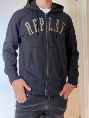  Replay hoodie  - Säljer en svart hoodie från Replay med dragkedja och stor REPLAY-text broderad över bröstet. Hoodien har två fickor framtill, ribbade muddar och en klassisk huva med dragsko. Perfekt för en chill och avslappnad stil.