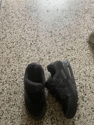 Svarta Nike Air Max sneakers - Säljer ett par helsvarta Nike Air Max sneakers med klassisk chunky sula och synlig Air-enhet. Skorna har mesh och syntetdetaljer för en sportig look och är perfekta för dig som gillar streetwear och komfort. Swoosh-loggan syns på sidan.