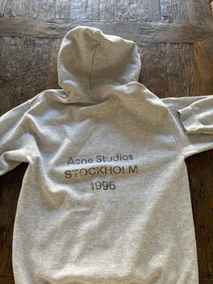 Grå hoodie Acne Studios Stockholm - Snygg grå hoodie från Acne Studios med tryck på ryggen där det står 'Acne Studios STOCKHOLM 1996'. Klassisk passform med huva och mjukt material. Perfekt för en avslappnad och trendig look.