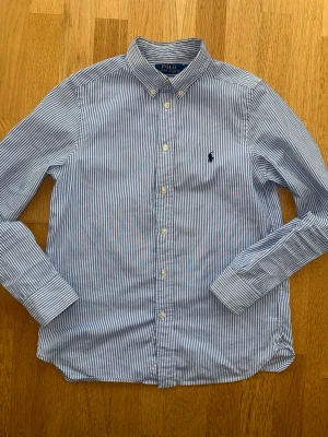 Randig skjorta från Polo Ralph Lauren - Klassisk blåvit randig skjorta från Polo Ralph Lauren med button-down krage och broderad logga på bröstet. Skjortan har långa ärmar och vita knappar framtill. Perfekt för en stilren och tidlös look.