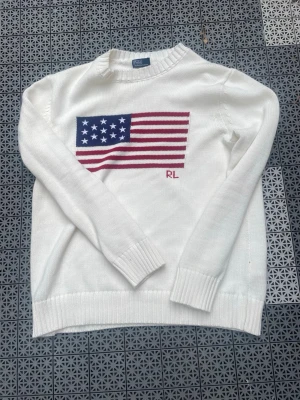 Vit stickad tröja Polo Ralph Lauren - Vit stickad tröja från Polo Ralph Lauren med amerikansk flagga och RL-brodyr på bröstet. Tröjan har rund halsringning, ribbade muddar och nederkant. Perfekt för dig som gillar klassisk och stilren design. Passar dig som är runt 180, Inga som helst defekter 
