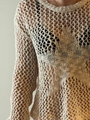 Beige stickad meshtröja med stjärna - Säljer en beige stickad meshtröja med lång ärm och en stor vit stjärna framtill. Tröjan har ett luftigt nätmönster och rund halsringning. Perfekt för lager på lager och en cool, avslappnad stil.