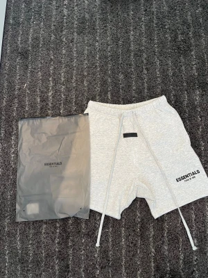 Grå Essentials shorts med snörning - Säljer ett par light oat shorts från Essentials med tryck på vänster ben. Shortsen har elastisk midja med långa snören och två sidofickor. Perfekta för chill dagar eller träning. Kommer med originalpåse. 