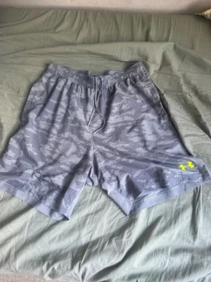 Grå camo shorts från Under Armour - Snygga grå shorts med camomönster från Under Armour. De har elastisk midja med dragsko och en gul logga på vänster ben. Perfekta för träning eller chill. Tillverkade i ett lätt och snabbtorkande material.